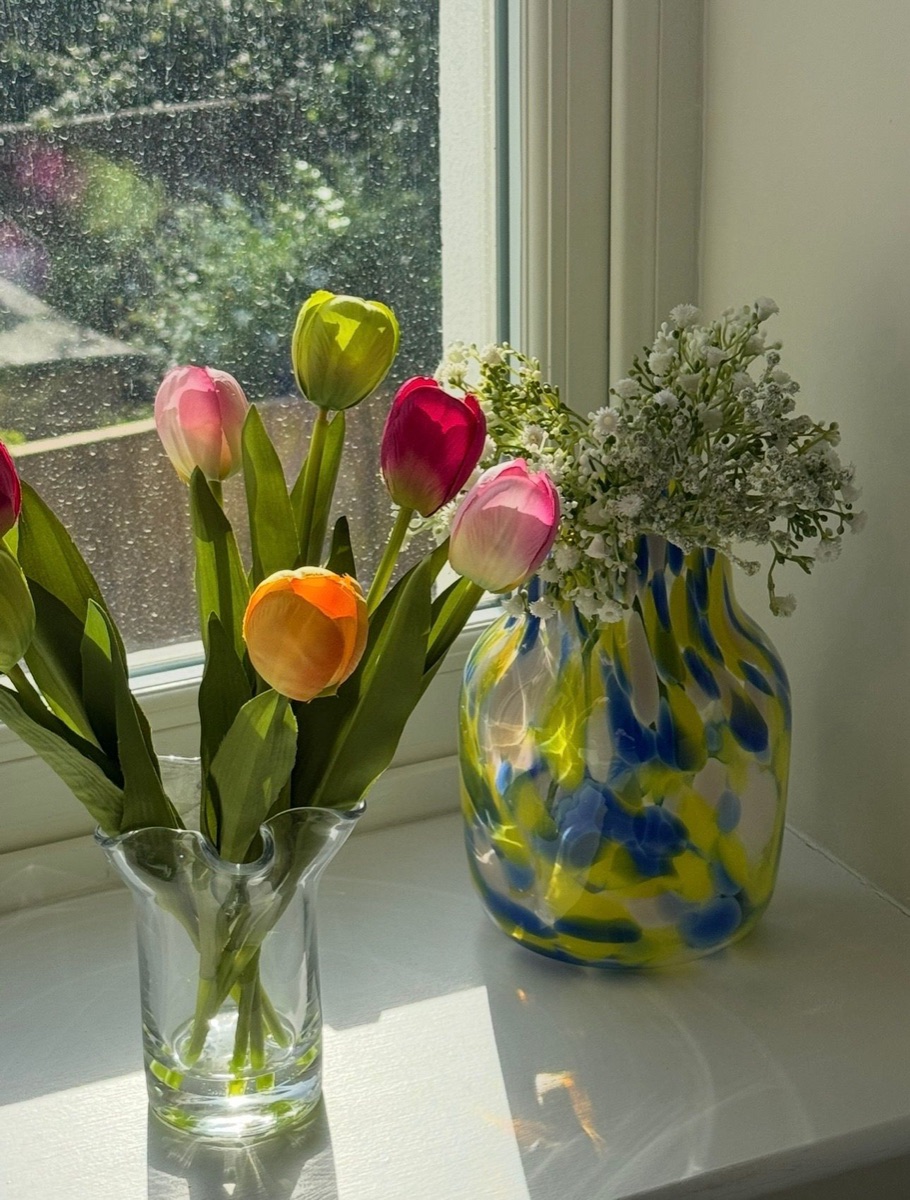 Pink and orange tulips beside a blue and yellow glass vase on a sunny windowsill
